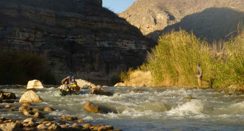 two people paddle a canoe through whitewater while high canyon walls stand in the background